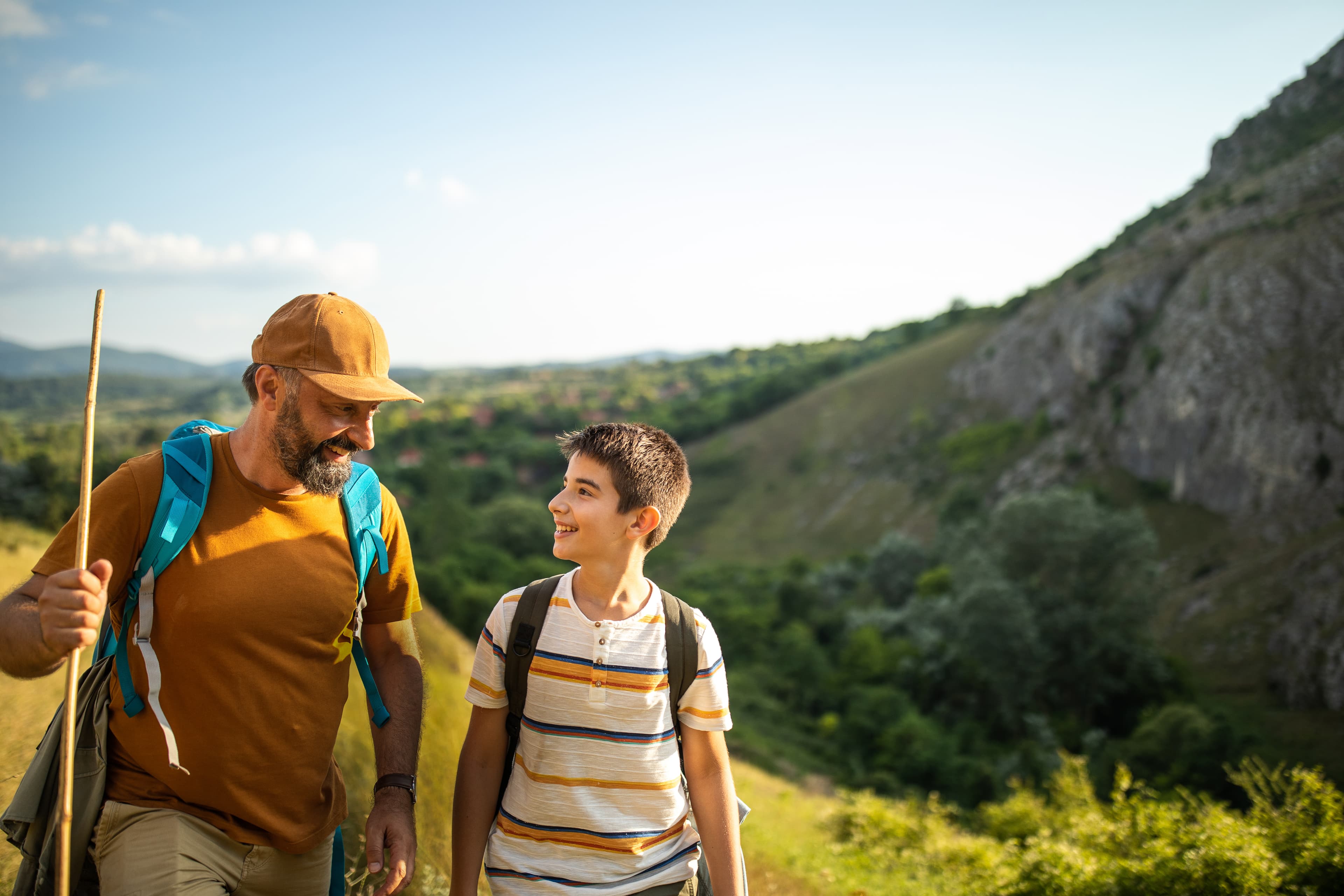 Man and boy with backpacks smiling at each other on a sunny hillside hike